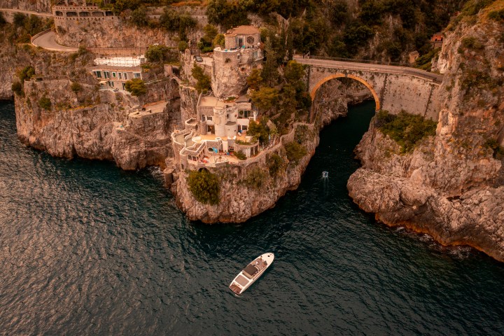 Aerial view of coastal cliffs with houses and arch bridge, boat on water.