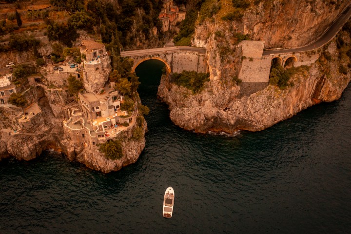 Coastal cliff with villas, arched bridge, and boat on dark water at sunset.