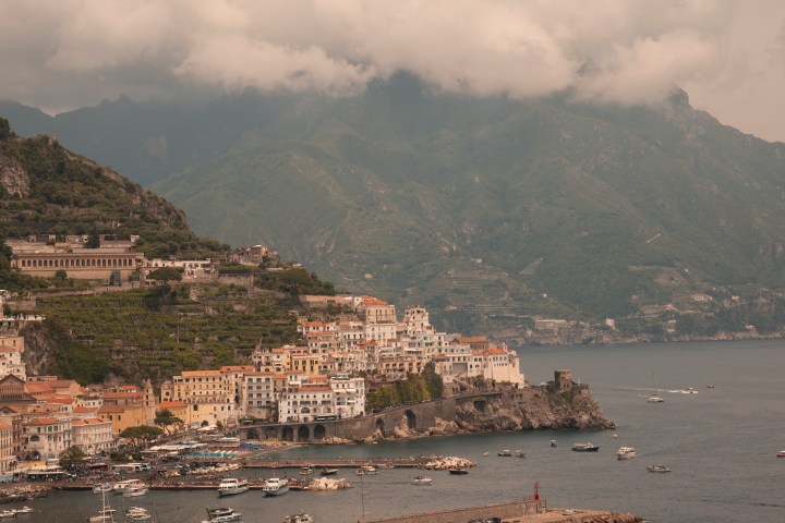Coastal town with colorful buildings, boats, and mountains under cloudy sky.