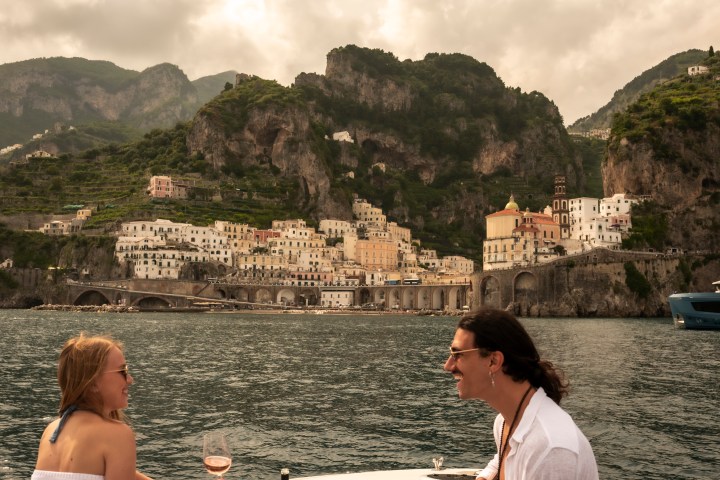 Two people enjoying a boat picnic with wine, near a coastal town with mountains in the background.