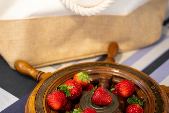 Strawberries in a wooden ship wheel bowl beside a canvas bag with 'Designers of Charming Cruises' logo.
