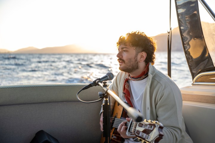 Person playing guitar on a boat at sunset with mountains in the background.