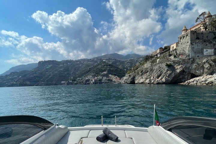 View from a boat with white cushions, drinks, and coastline in the background under a blue sky.