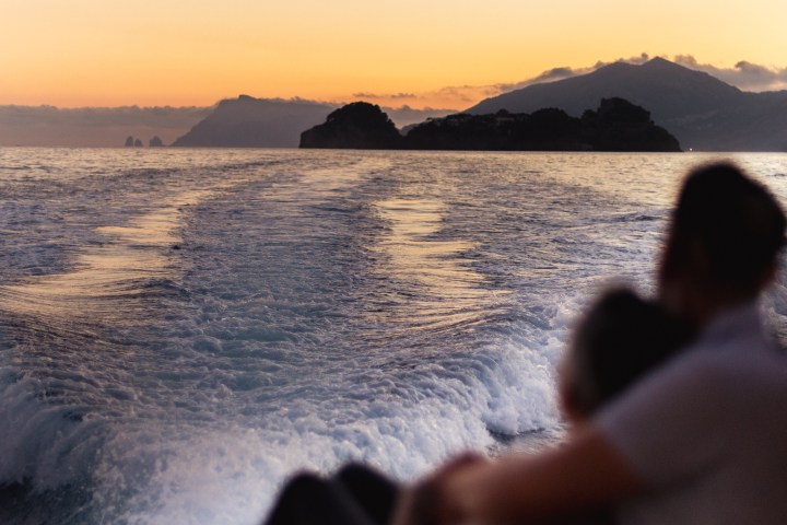 Couple on boat at sunset with island silhouette and ocean waves.