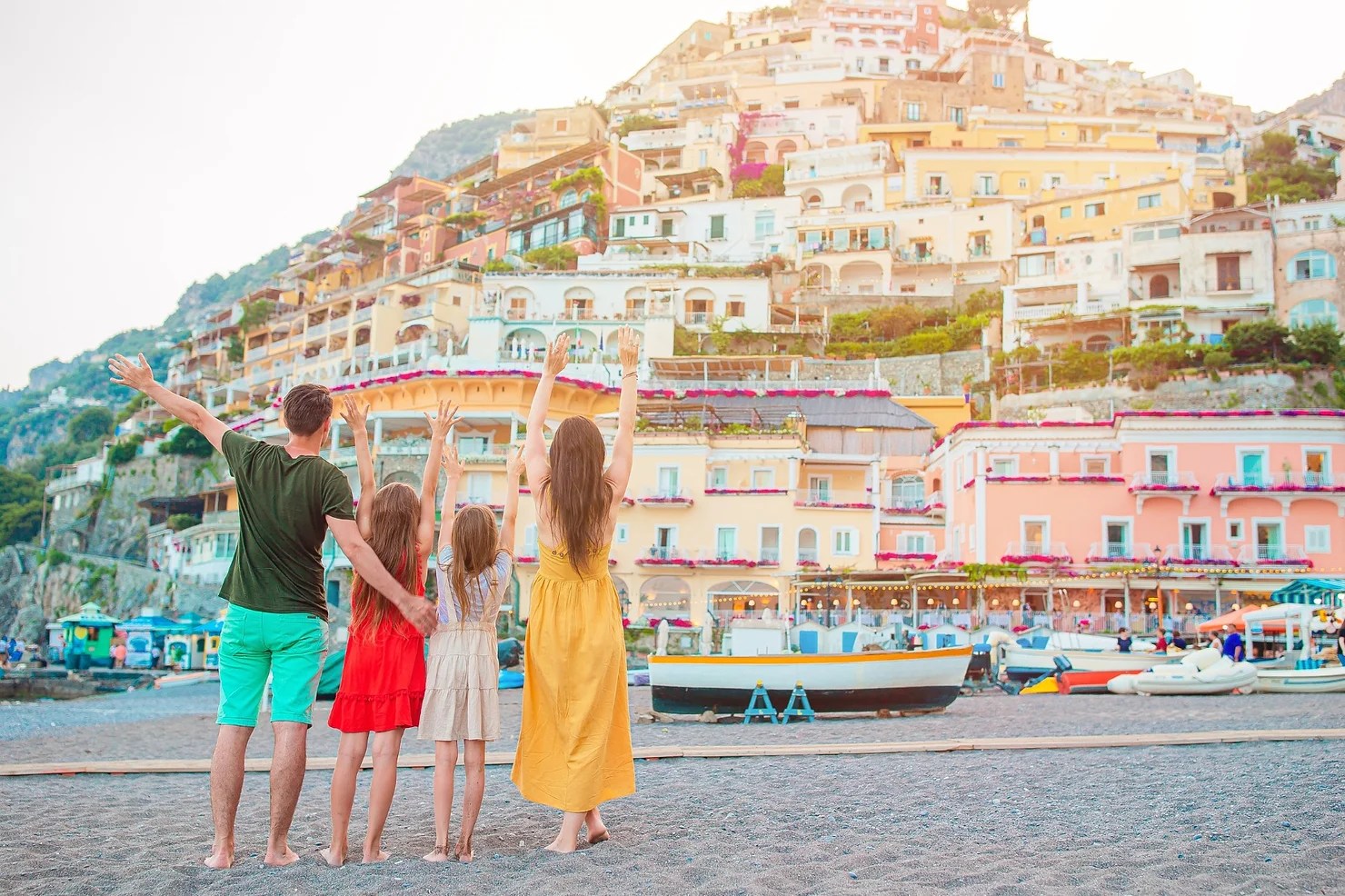 Family of four with raised arms, facing colorful hillside buildings on a beach.