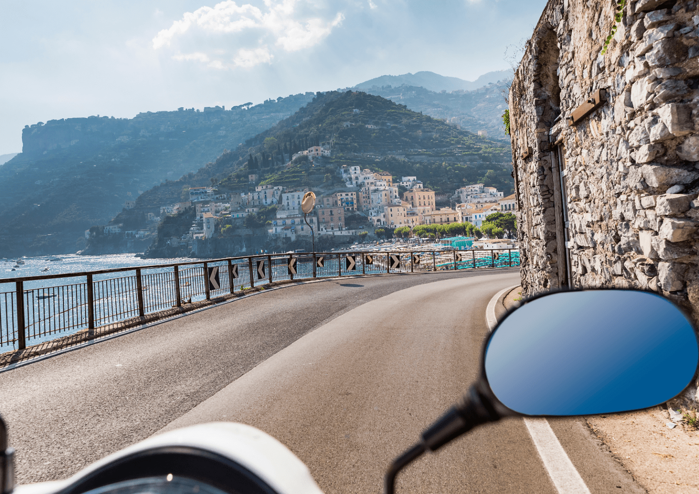 Coastal road with a scooter mirror, village on hillside, and blue sea on a sunny day.