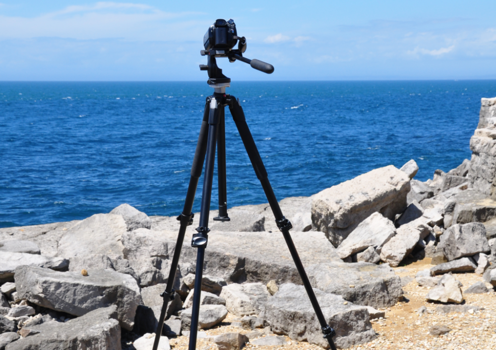 Camera on a tripod by a rocky ocean shore under a clear blue sky.