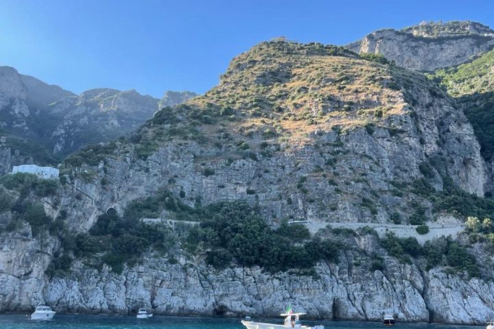Boats on turquoise water beneath rocky cliffs with sparse vegetation under a clear blue sky.
