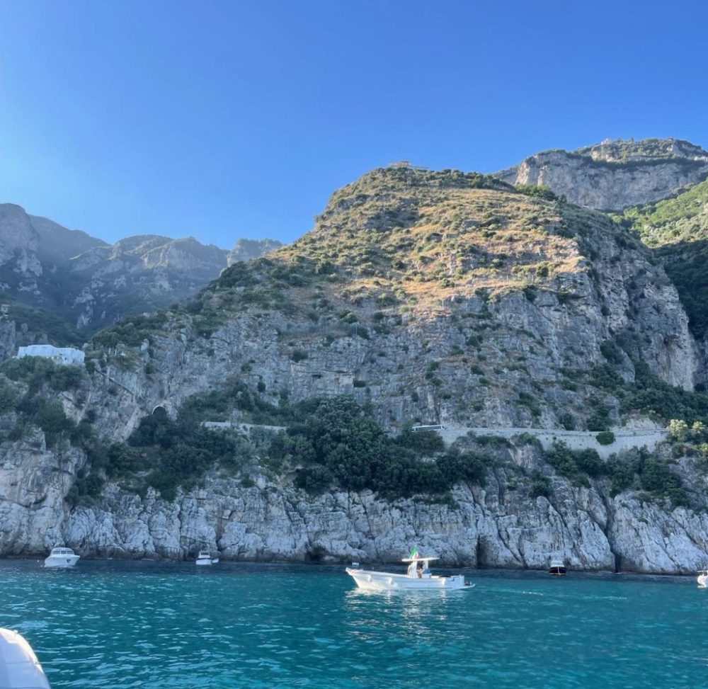 Boats on turquoise water beneath rocky cliffs with sparse vegetation under a clear blue sky.