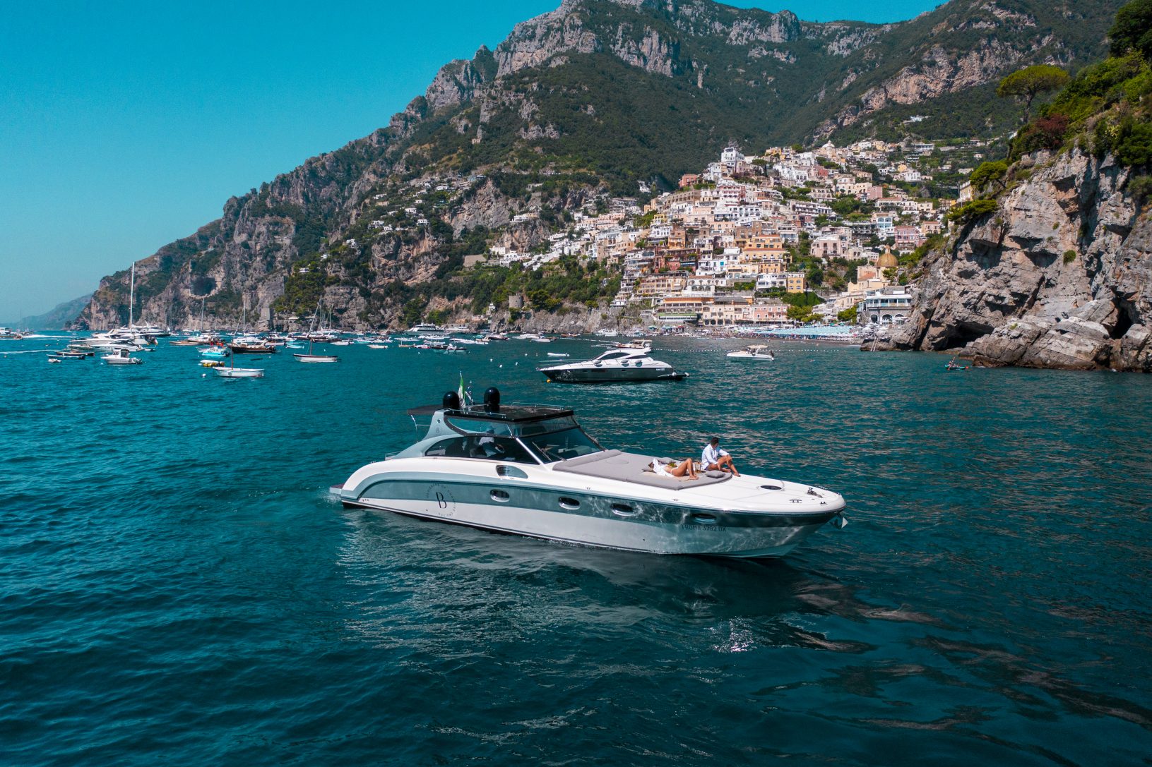 Luxury yacht on blue water with coastal town and rocky cliffs in background under clear sky.