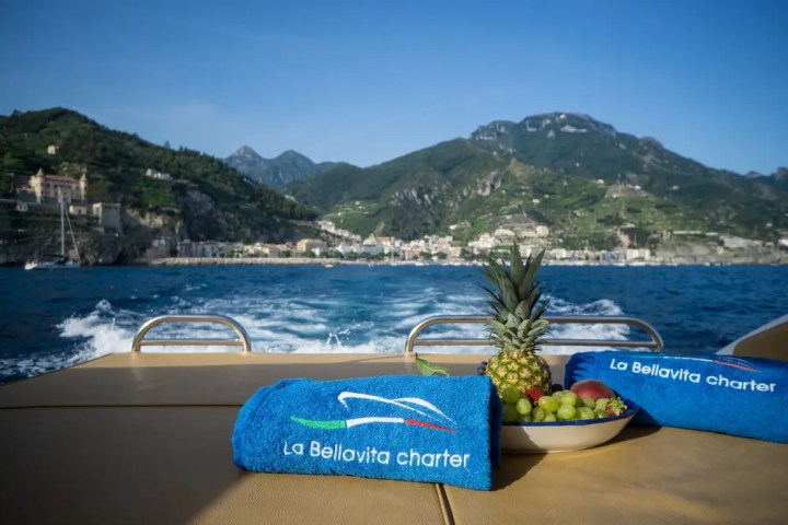 Boat deck with blue towels, fruit bowl, and coastal mountains in the background.