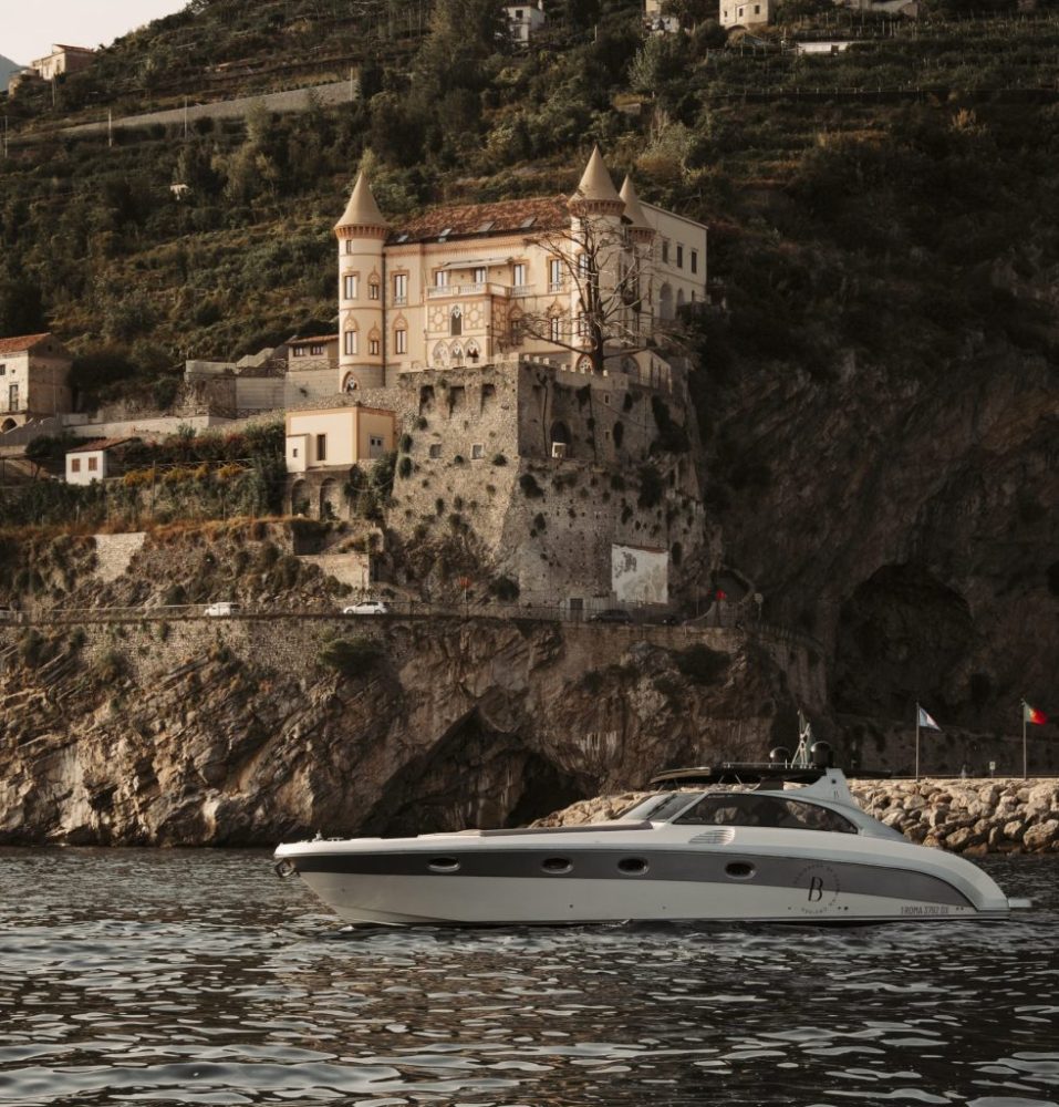 Luxury yacht in front of a cliffside castle with turrets, surrounded by lush greenery.