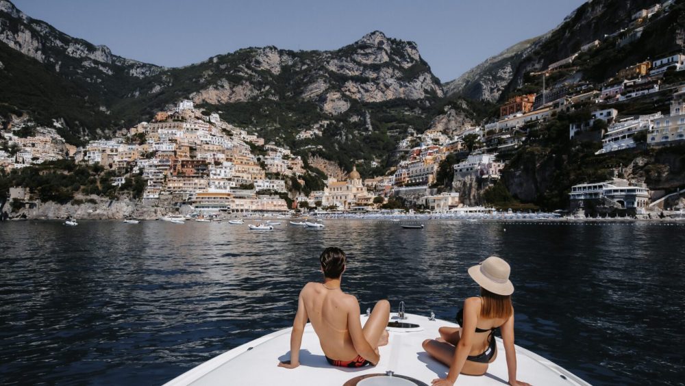 Two people relaxing on a yacht in front of a coastal town with mountains in the background.