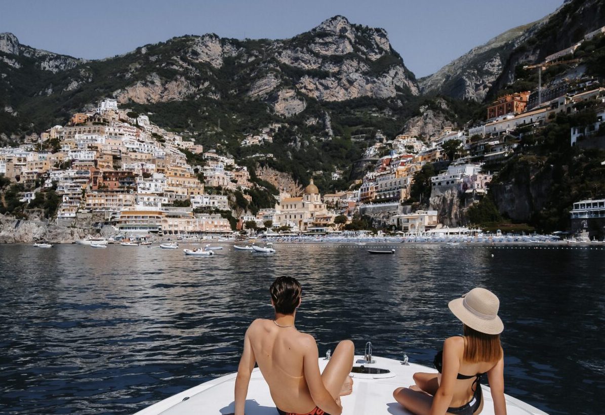Two people relaxing on a yacht in front of a coastal town with mountains in the background.
