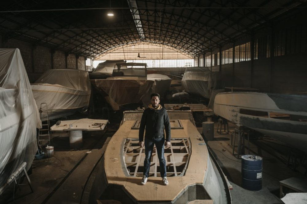 Person standing on an unfinished boat hull in a dimly lit workshop with boats covered in plastic.