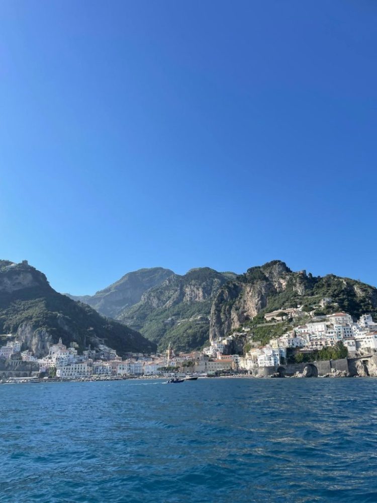 Coastal town with white buildings, mountains, and blue sea under a clear sky.