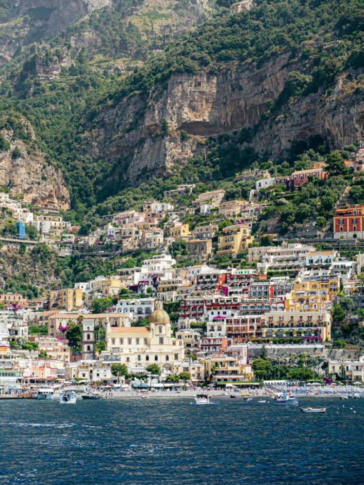 Colorful hillside village with church dome by the sea, surrounded by cliffs and greenery.
