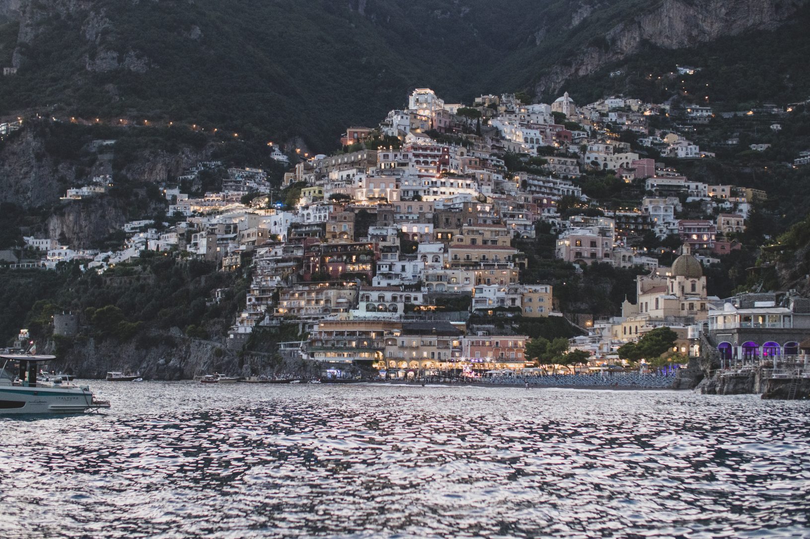 Hillside town with colorful buildings lit up at night, viewed from the water, with a small boat in foreground.