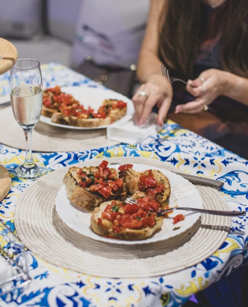 Bruschetta on a patterned tablecloth with a glass of champagne and person holding fork.
