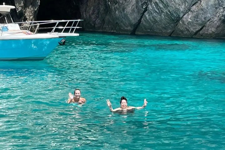 Two people swim in clear turquoise water near a rocky cave, with part of a boat visible.