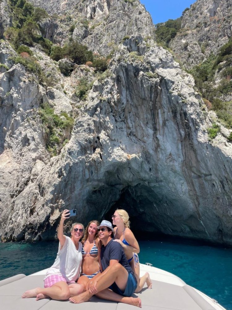 Four people take a selfie on a boat near rocky cliffs and a cave.