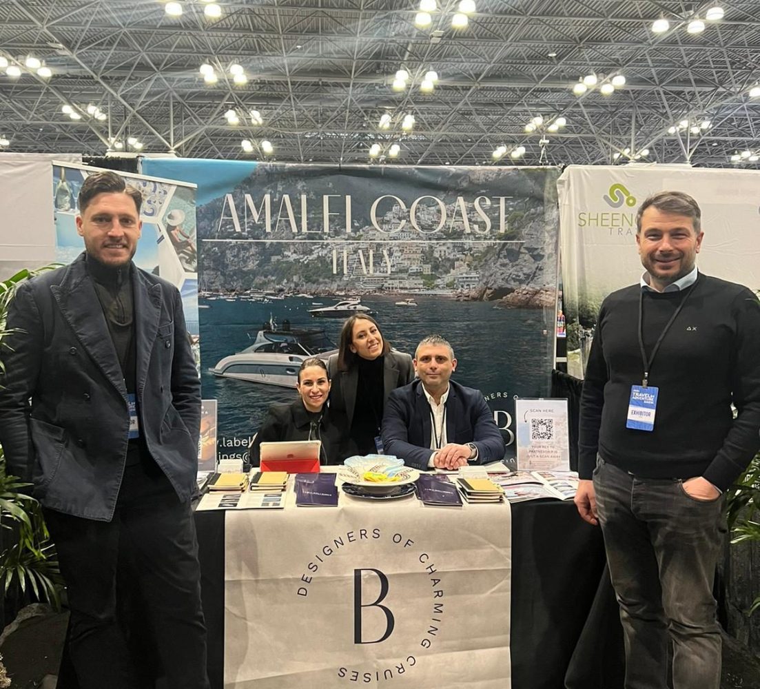 Five people smiling at a booth with 'Amalfi Coast Italy' banner in a convention center.