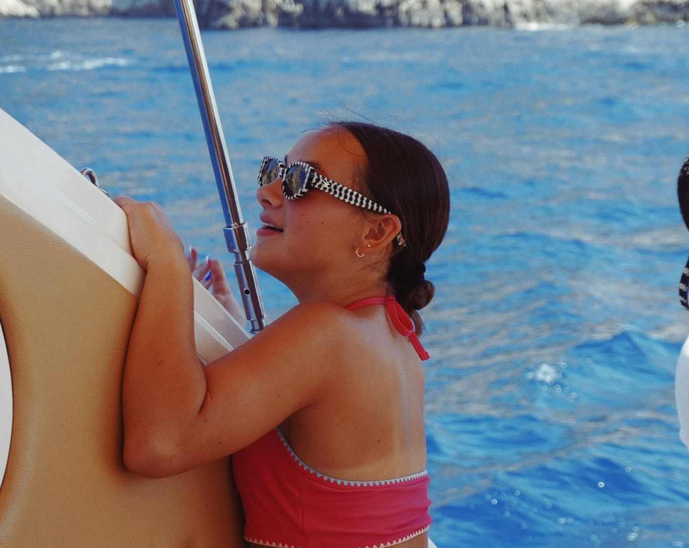 Girl with sunglasses on a boat, holding a railing with blue sea in background.