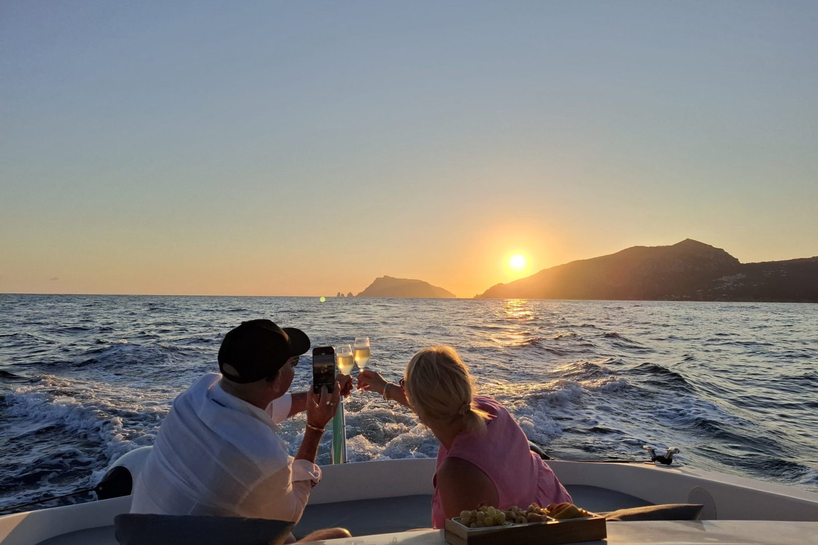Two people on a boat toast with champagne at sunset with an island in the background.