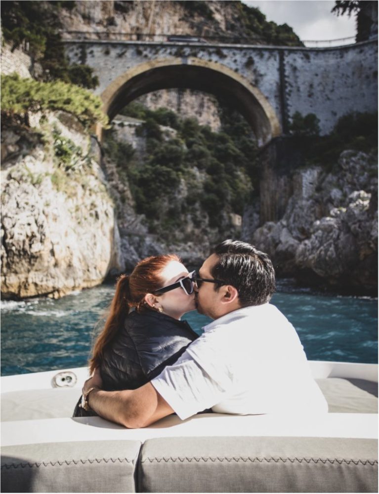 Couple kissing on a boat with a stone bridge and cliffs in the background.