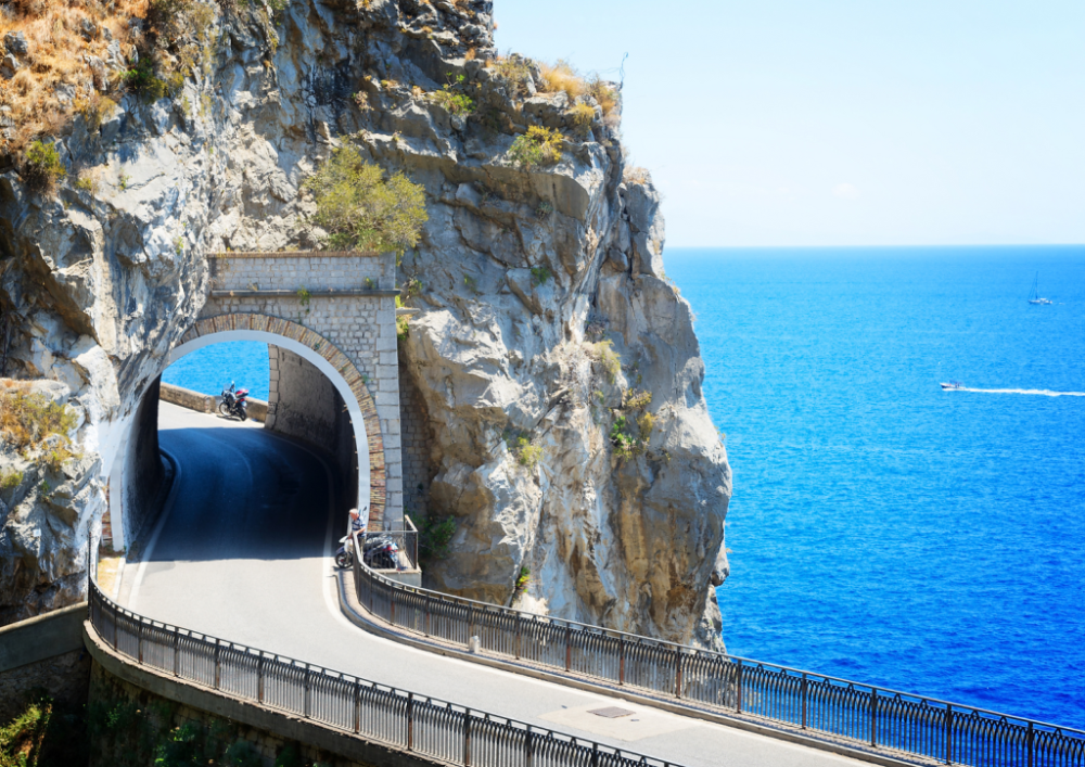 Coastal road tunnel with motorcyclist, cliffside and ocean in background on a sunny day.