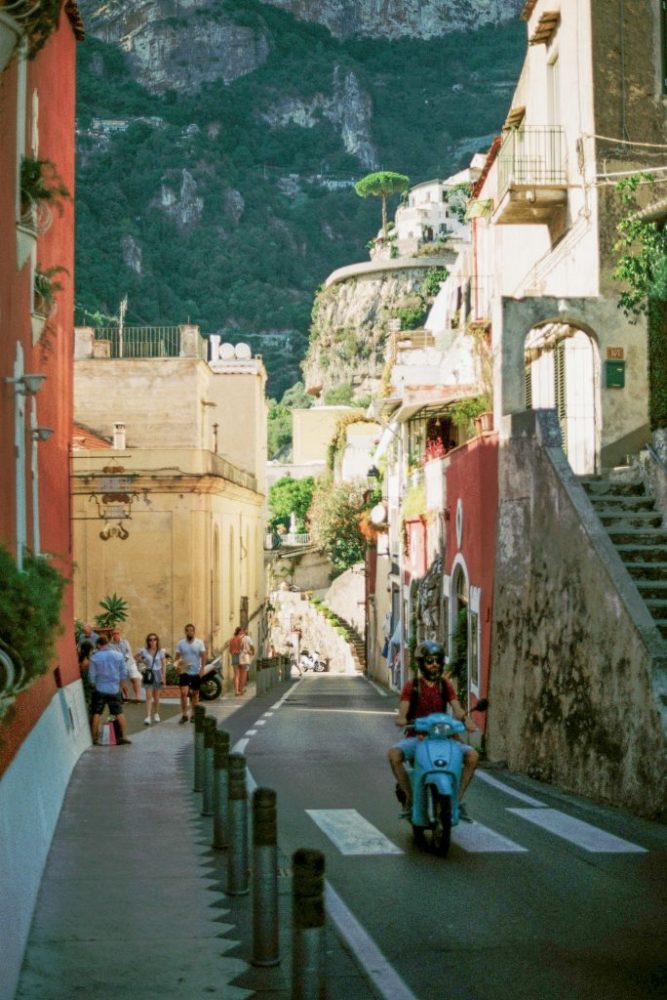 Narrow Italian street with a person riding a scooter and pedestrians, mountain in background.