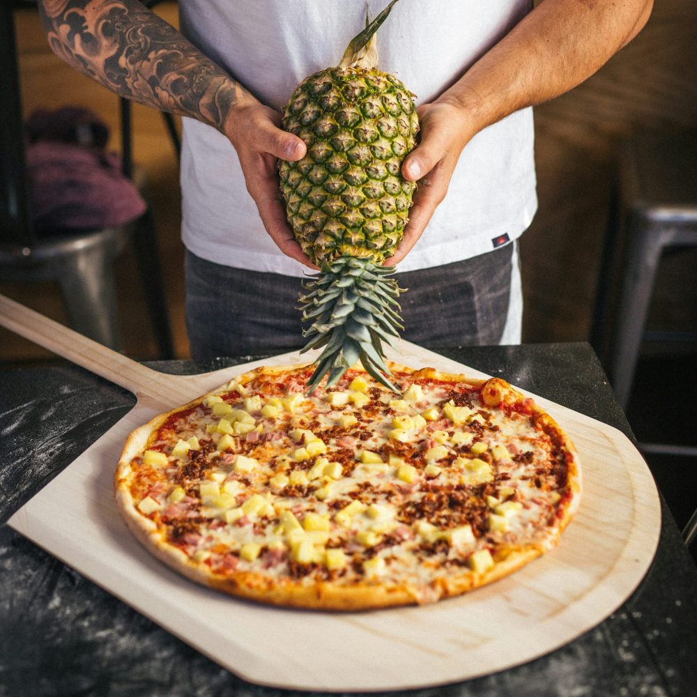 Person holding pineapple above a pizza with pineapple and ham on a wooden board.