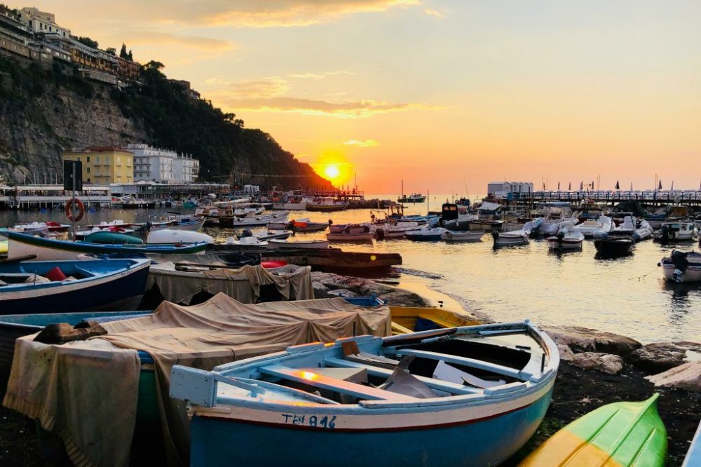 Boats in a harbor at sunset with cliffs and buildings in the background.
