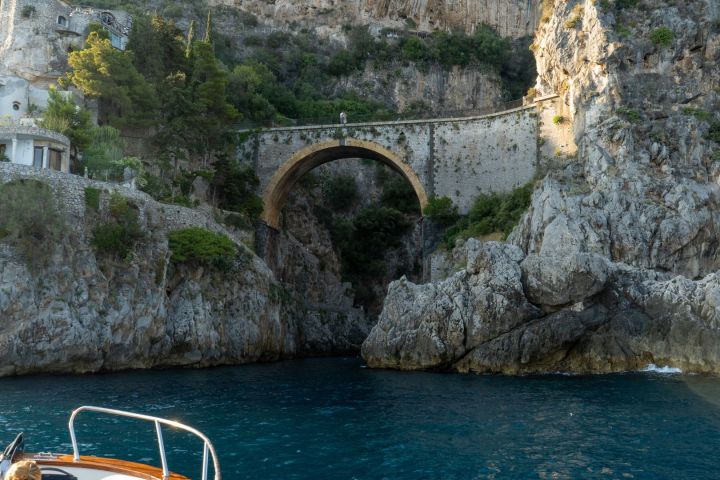 Boat in blue water near rocky cliffs with an arch bridge above.