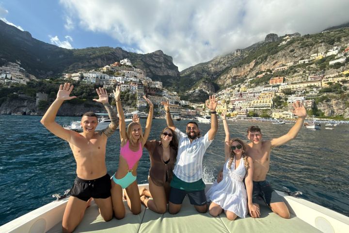 Six smiling people on a boat with Positano's colorful cliffside buildings in the background.