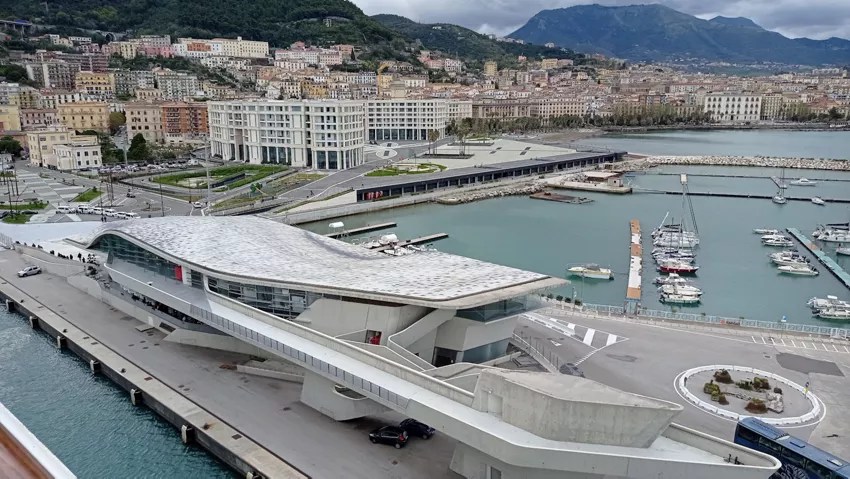 Modern waterfront building with curved roof, marina with boats, and hilly cityscape in background.