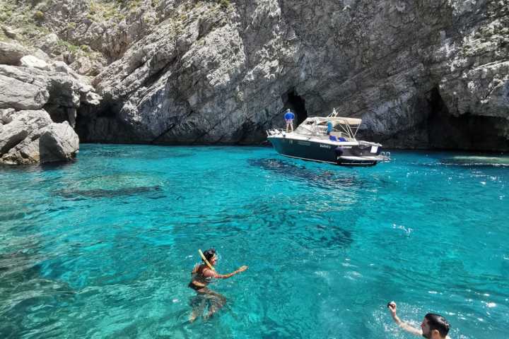 Two people swimming in clear blue water near rocky cliffs and a boat.