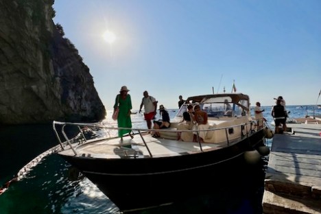 People on a boat docked near a rocky cliff with the sun shining brightly.