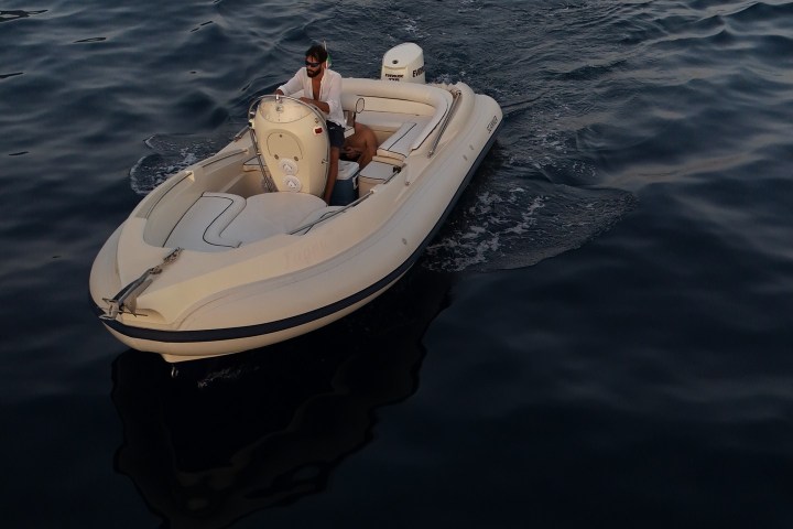 Person driving a small boat on calm, dark waters near a rocky coastline.