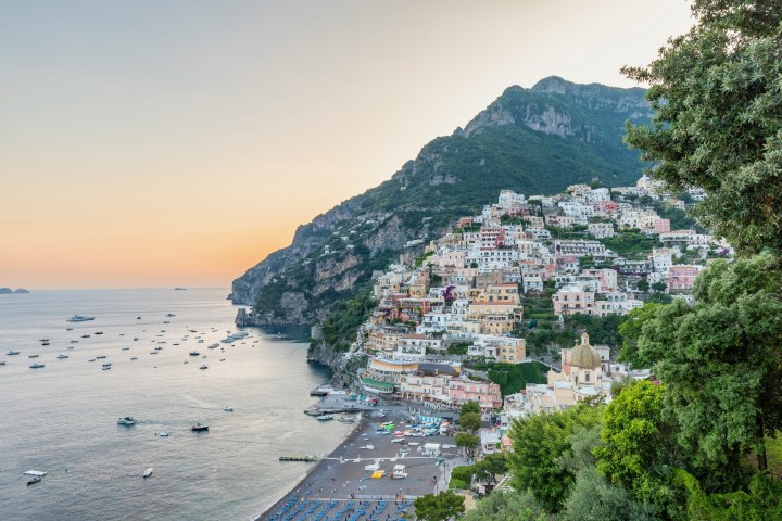 Sunset view of a coastal town with colorful hillside buildings and boats on calm water.
